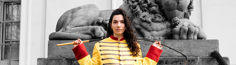 Eine junge Frau blickt mit einem neutralen Gesichtsausdruck in die Kamera. Sie trägt eine gelbe Dompteurjacke und hat eine Zirkuspeitsche über ihre Schultern gelegt. Sie steht vor der Statue eines sitzenden Löwen. Stückbild des Stückes Iphigenie