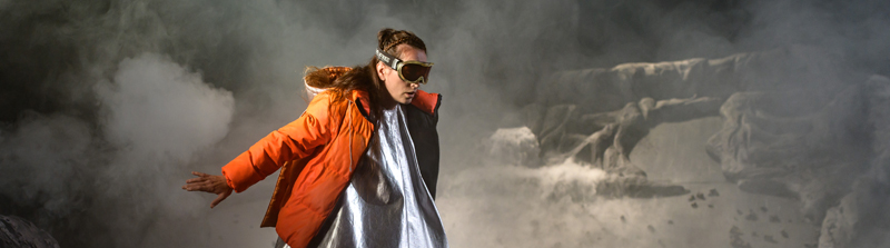 Szenenfoto der Inszenierung »Stella«: Eine junge Frau in einer orangefarbenen Ski-Jacke, einem silberfarbenen Kleid und mit einer Ski-Brille steht mit nach hintem gestreckten Arm vor einer Felslandschaft im Nebel. Stückbild des Stückes Stella