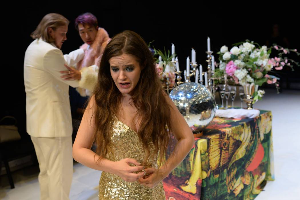 Das Bild zeigt ein Szenenfoto aus »Euridice«. Im Vordergrund steht eine Frau mit langen braunen Haaren. Sie trägt ein goldenes Glitzerkleid, hat silber lackierte Fingernägel und singt gerade. Im Hintergrund steht ein Tisch mit glitzernder Diskokugel, weißen Kerzen und rosa- und weißen Blumen. Es sind außerdem zwei Männer zu sehen, die sich gegenüberstehen und singen. Ein Mann hat schulterlange dunkelblonde Haare und trägt einen weißen Anzug. Der andere Mann trägt einen Pelzmantel in den Farben rosa- und creme. Er hat schwarze kurze Haare, die am Pony lila gefärbt sind. © Candy Welz