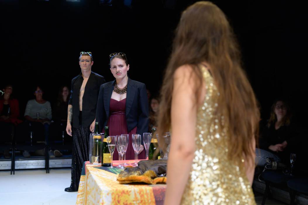 Das Bild zeit ein Szenenfoto aus »Euridice«. Im Vordergrund steht eine Frau mit dem Rücken zur Kamera an einem Tischende. Sie hat lange braune Haare und trägt ein goldenes Glitzerkleid. Ein Mann und eine Frau stehen am anderen Ende des Tisches. Auf dem Tisch stehen Sektflaschen und Sektgläser. Der Mann und die Frau tragen beide verspiegelte Sonnenbrillen. Die Frau hat ein weinrotes Kleid und trägt eine dreilagige braune Kette mit kleinen Perlen. Sie trägt einen Schnauzbart und schaut skeptisch zur Frau am anderen Ende des Tisches. Der Mann trägt einen schwarzen Anzug mit glitzernder Hose. An einem Arm trägt er Tattoos. Auch der Mann blickt skeptisch zur Frau am anderen Ende des Tisches. © Candy Welz