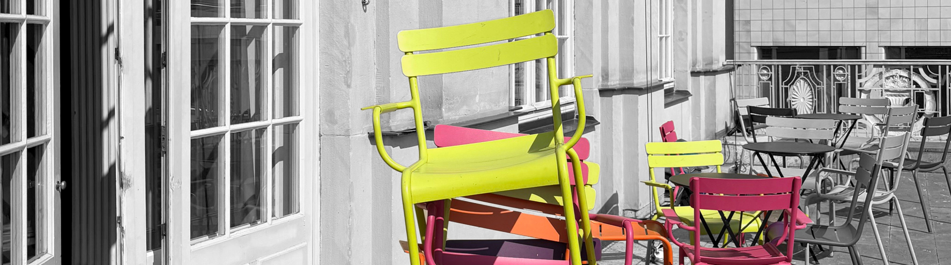 Zu sehen ist ein Stapel neonfarbener Stühle rechts vor einer halb geöffneten Tür mit Glasscheiben sowie weitere Stühle, die um Tische herum stehen.