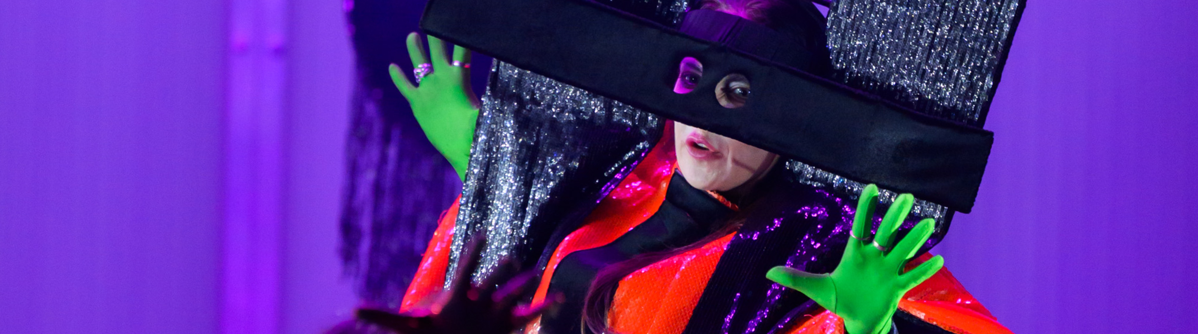 Das Bild zeigt eine kostümierte und maskierte Frau vor einem lilafarbenen Hintergrund.