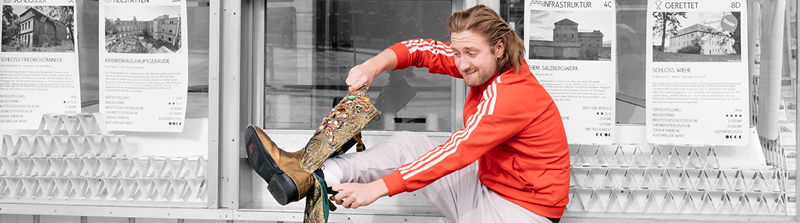 Ein Mann mit schulterlangen Haaren und Bart hebt sein rechtes Bein und versucht mit beiden Händen einen goldenen Stiefel anzuziehen. Der Stiefel ist mit farbigen Steinen besetzt. Der Mann beißt sich auf die Unterlippe und runzelt die Stirn. Er trägt eine orange-farbene Jacke mit weißen Streifen. Der Hintergrund ist schwarz-weiß. Stückbild des Stückes Felix Krull