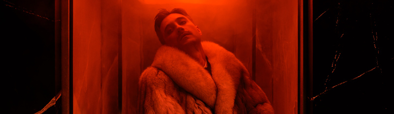 Szenenfoto der Inszenierung »Felix Krull«: Ein Mann in einem Pelzmantel steht in einem rot ausgeleuchteten vernebelten engen Raum. Stückbild des Stückes Felix Krull