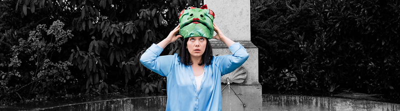 Eine Frau in einem blauen Hemd schaut verwundert nach oben. Auf ihrem Kopf sitzt ein grüner Hut, der die Form eines Frosch-Gesichts hat. Sie hält ihn mit beiden Händen fest. Der Hintergrund ist schwarz-weiß. Stückbild des Stückes Gullivers Reisen