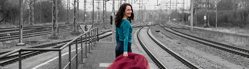 Eine Frau in blauem Oberteil steht neben Bahngleisen. Sie hat den Rücken zur Kamera gedreht und blickt über die rechte Schulter nach hinten. Sie grinst in die Kamera. In ihrer rechten Hand hält sie einen weinroten Tüll-Rock. Der Hintergrund des Bildes ist schwarz-weiß. Stückbild des Stückes Kairos