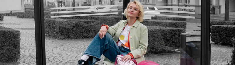 Eine Frau mit blonden Haaren schaut in die Kamera. Sie sitzt an einer Bushaltestelle und ihr rechtes Bein steht auf der Bank. Ihre rechte Hand stützt sie auf ihrem Knie ab. Sie trägt einen mintgrünen Mantel, Jeans und eine bunt gepunktete Bluse. Der Hintergrund ist schwarz-weiß. Stückbild des Stückes Draußen vor der Tür