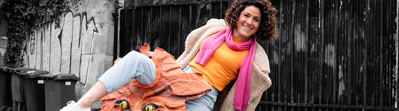 Eine Frau mit braunen Locken schaut lachend in die Kamera. Sie trägt ein orange-farbenes T-Shirt und einen pinken Schal. Sie hat ihr rechtes Bein gehoben und eine rote Fuchsmaske aus Pappmaché liegt um ihr Bein. Stückbild des Stückes Rechnitz (Der Würgeengel)