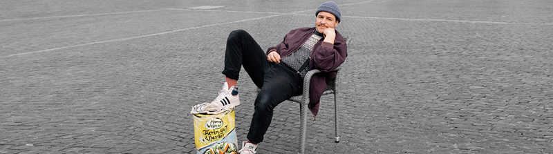 Ein Mann mit einem blauen Beanie und einer dunkellila Jacke sitzt draußen auf einem Stuhl. Er hat sich zurückgelehnt und sein rechtes Bein steht auf einer vergrößerten Nachahmung einer "Thüringer Allerlei" Dose. Er hat sein Gesicht auf seinen linken Arm gestützt und schaut entspannt in die Kamera. Stückbild des Stückes Maß für Maß