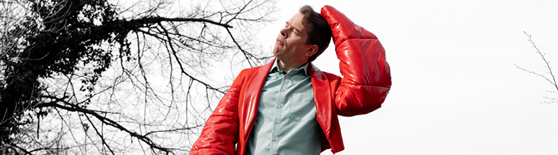 Ein Mann mittleren Alters trägt eine knallrote Jacke aus Fake-Leder. Die Arme der Jacke sind geformt wie Krabbenarme. Mit der linken Hand kratzt er sich am Kopf, seine Augen sind geschlossen und er hat einen nachdenklichen Gesichtsausdruck aufgesetzt. Der Hintergrund des Bildes ist schwarz-weiß, zu sehen ist nur der Himmel und ein paar Äste. Stückbild des Stückes Heute Nacht oder nie