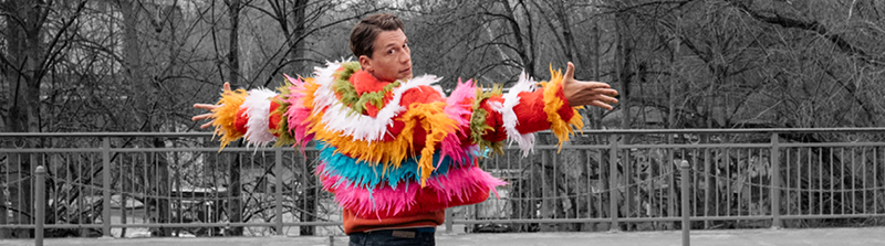 Ein Mann steht mit dem Rücken zur Kamera auf einer Brücke. Er hat beide Arme zur Seite gestreckt und trägt eine bunte Jacke, die mit Federn besetzt ist. Seinen Kopf hat er nach hinten zur Kamera gedreht, in die er hineinschaut. Der Hintergrund ist schwarz-weiß. Stückbild des Stückes Kiss me, Kate
