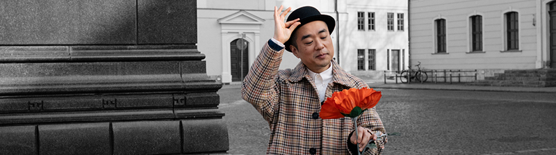 Ein Mann in einem gemusterten Mantel schaut mit einem freundlichen Gesichtsausdruck auf eine große orange-farbene Blume in seiner linken Hand. Die rechte Hand hat er gehoben und berührt damit die Krempe seines schwarzen Hutes. Stückbild des Stückes Werther