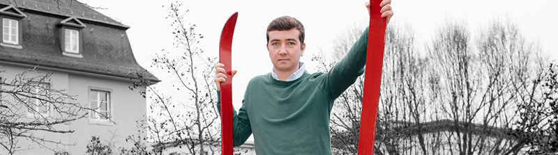 Ein Mann in grünem Pullover schaut mit neutralem Gesichtsausdruck in die Kamera. In beiden Händen hält er rote Skier. Der Hintergrund ist schwarz-weiß. Stückbild des Stückes Der Barbier von Sevilla