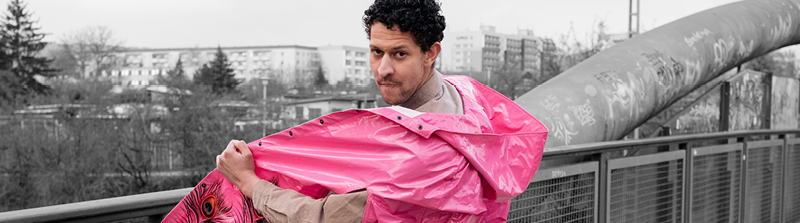 Ein Mann steht auf einer Brücke. Er hat seinen Körper halb abgewandt, schaut aber über seine linke Schulter in die Kamera. Er trägt einen pinken Regenmantel, den er mit der linken Hand offen hält. Der Hintergrund ist schwarz-weiß. Stückbild des Stückes Der Mordfall Halit Yozgat